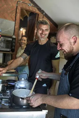 two men in a kitchen preparing food in a pan at HOSTEL CASA C Alojamiento, Coworking y Centro Cultural in Villa General Belgrano