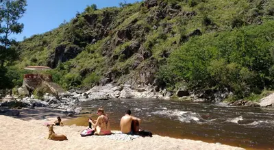 a group of people sitting on a beach next to a river at Departamento Valle de Luz in Alta Gracia
