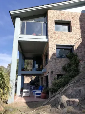 a large stone house with glass windows and blue chairs at casa del lago -villa carlos paz in Villa Carlos Paz