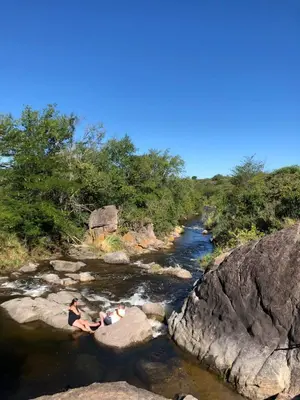 two people sitting on rocks in a river at CABAÑAS FENIX con EL ARROYO AMBOY RODEANDO LA PROPIEDAD reservar con 24 hs de anticipación in Villa Amancay
